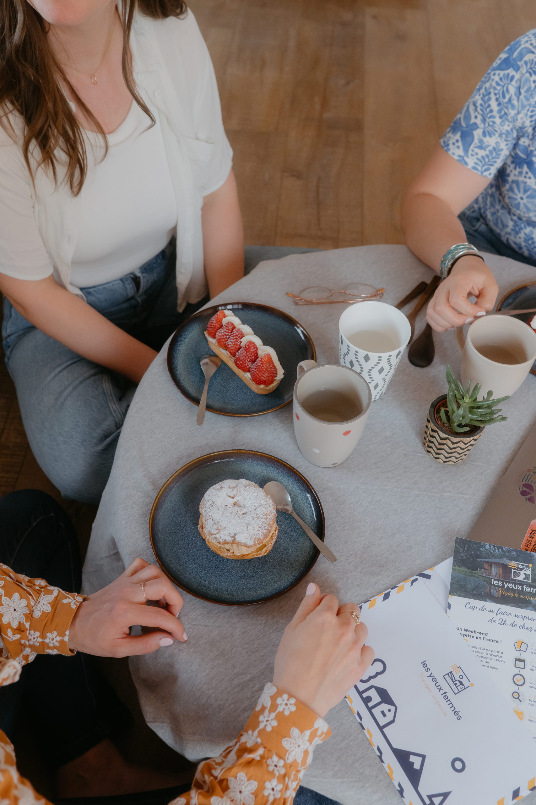 Mathilde, Sarah et Marlène sont assises autour d'une table avec des pâtisseries, des tasses et ainsi que des enveloppes surprises et un PC pour travailler.
